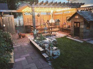 Cozy backyard patio with lights, a wooden playhouse, and a dog resting on the brick path.